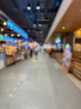 Blurred image of people walking down a wide, well-lit corridor in a modern shopping mall or large retail center, with colorful bokeh lights.