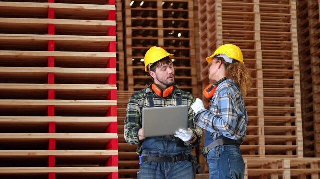 Professional carpenter and engineer wearing safety helmets inspect stacked timber inside a woodworking factory while using a laptop for production planning and quality control.