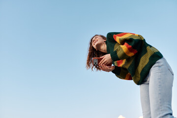 Happy young woman in colorful striped sweater laughing while holding smartphone against blue sky....