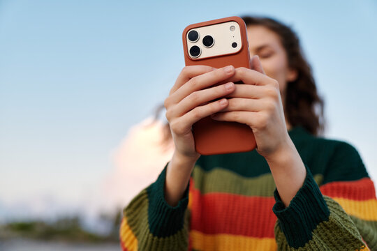 Young woman in colorful striped sweater holds smartphone taking photo outdoors, face hidden behind phone, blue sky background. Concept of mobile photography and social media lifestyle.