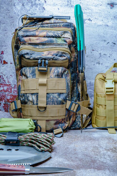 Camouflage tactical backpack and essential survival gear, including knives, paracord, and a saw, prepared for bushcraft adventures