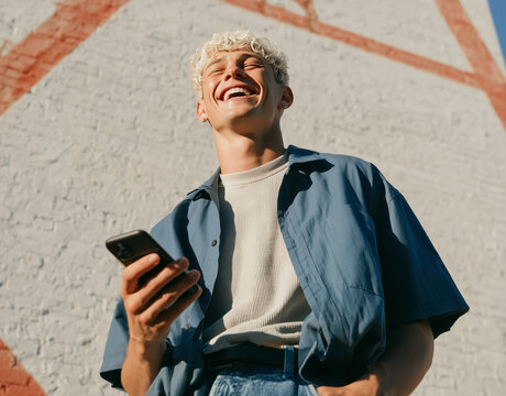 Young man laughs joyfully outdoors, holding phone. Sunlit wall with bold red stripes adds urban texture. Casual denim shirt, knitted turtleneck beneath. Radiant smile suggests contentment or amusement