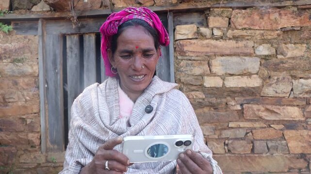 A rural woman is looking at a modern smartphone while sitting against a stone wall.
