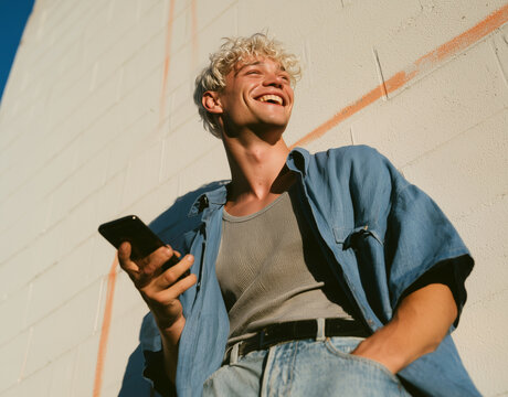 Young man laughs while holding a smartphone. He wears a blue shirt, jeans, and a patterned tee. Sunlight highlights his curly, blond hair and joyful expression