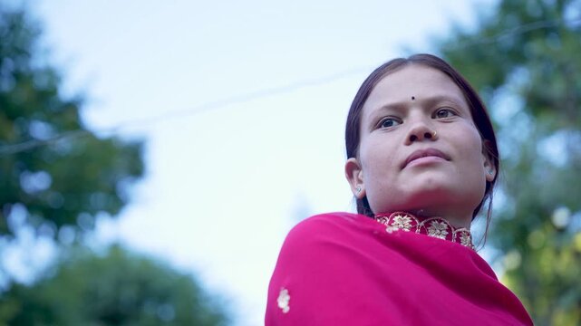 A beautiful local woman wearing a bright red shawl stands thoughtfully outdoors in the countryside.