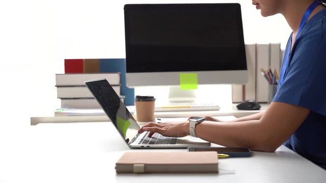 Medical Professional Working on Laptop: A medical professional, seen from the side, diligently types on a laptop at a desk, with a computer monitor and stacked books in the background.