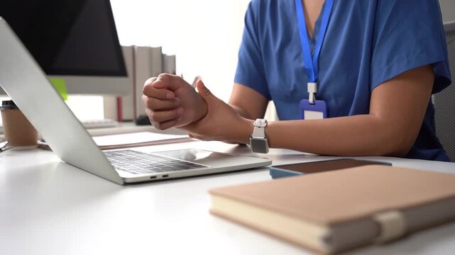 Medical Professional Working at Desk: A close-up shot shows a healthcare worker in blue scrubs sitting at a desk, their hands clasped in thought or concern as they look at a laptop.