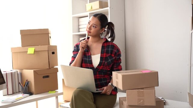Small business owner contemplating strategy: A young woman sits among stacks of cardboard boxes, laptop in front of her, deep in thought about her growing online retail business.
