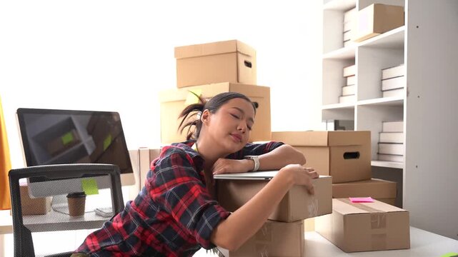 Overwhelmed Entrepreneur Slumbers: A young woman, looking exhausted, rests her head on a stack of boxes in her workspace, surrounded by shipping supplies and a computer.