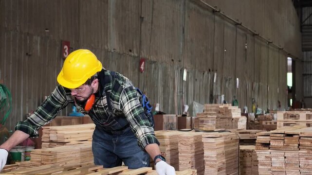 Carpenter wearing a hard hat, safety glasses, gloves, and hearing protection inspects wooden boards in a lumber factory workshop.