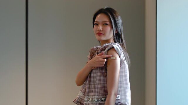 Happy young Asian woman pointing at bandage on arm after receiving vaccination or flu shot in clinic, promoting immunization and healthcare prevention for healthy lifestyle and immunity building.