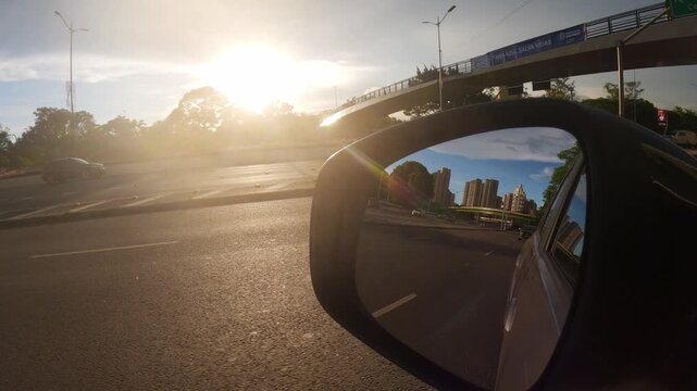 A view from a vehicle side mirror capturing the modern architecture of the Ibirapuera region during a golden hour sunset. The bright sun creates a dramatic lens flare against the urban traffic, showca