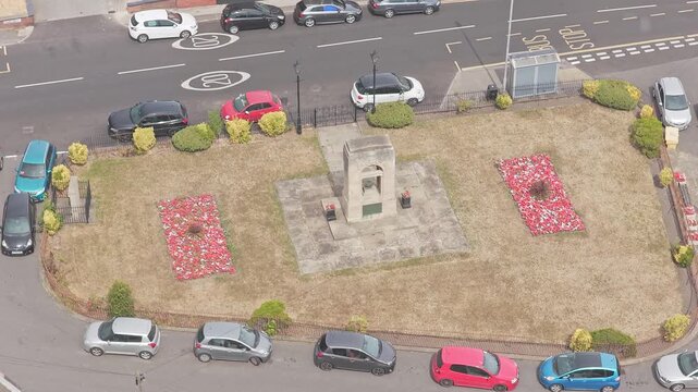 Greasbrough War Memorial stands between red poppy beds along Church Street, Greasbrough, Rotherham, South Yorkshire, England, with parked cars and surrounding streets visible, drone ascent