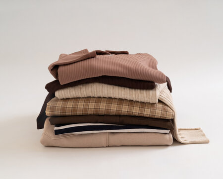 A neatly folded stack of cozy winter sweaters and casual shirts in neutral tones on a clean white background.
