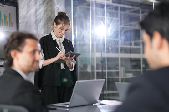 Focused businesswoman jots notes on a tablet with a stylus during a standing meeting as two out-of-focus managers watch from the foreground in a glass-walled coworking office.