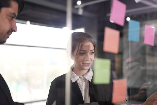 Soft smile from the young woman with a light brown bun as the bearded manager points out one of the sticky notes low on the transparent office wall between them.