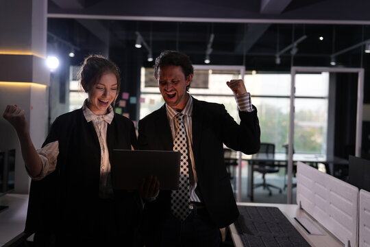 Screaming with joy at the laptop display, a young businesswoman and a bearded businessman celebrate an against the odds win during their long night shift at the office.