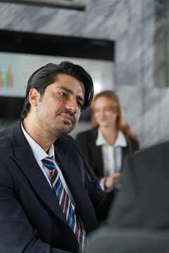 Businessman grimaces with visible frustration at the boardroom table while his ginger-haired colleague holds her smile in the soft background in a bright modern coworking office.
