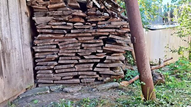 Stacked wooden boards stored beside a weathered shed in a rural backyard, surrounded by trees and vegetation. The scene shows aged timber, rustic construction materials, and a traditional outdoor sett