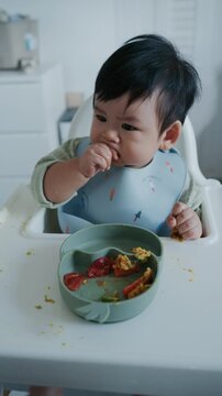 Asian baby boy eating boiled corn on high chair, BLW (baby-led weaning) concept, healthy solid food introducing to infant at home	
