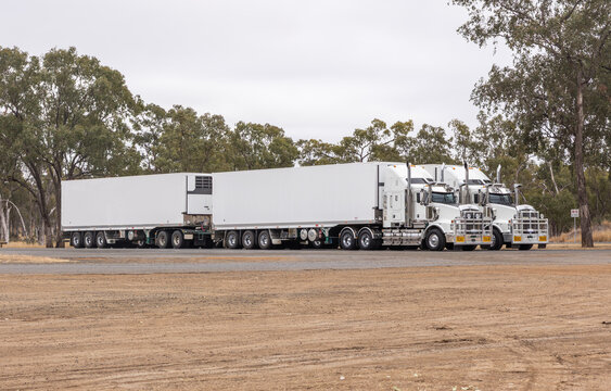 Two large, identical semi-trailer trucks, known as road trains, with refrigerated trailers, parked next to each other in the outback countryside under an overcast sky in central Queensland, Australia.