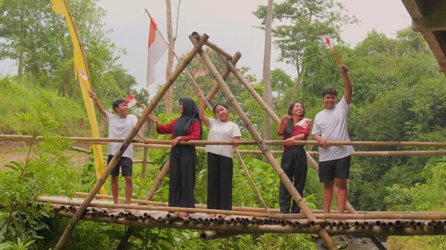 Five young Indonesian people standing on a wooden bridge while raising country flags with joyful expression. Indonesia Independence Day concept.