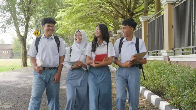 Four joyful young Asian students in school uniform are holding books and tablets while studying in the park.