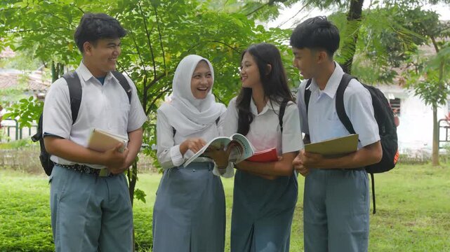 Four joyful young Asian students in school uniform are holding books and tablets while studying in the park.