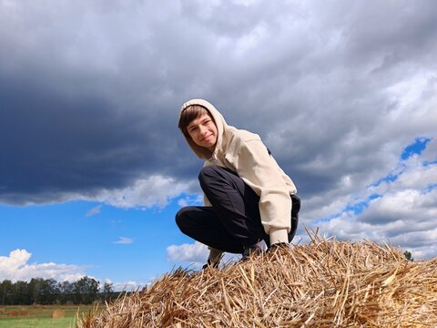 Boy Squatting on Hay Bale Under Stormy Sky. Teenage boy in beige hoodie squatting on a large hay bale against a dramatic cloudy sky. Rural outdoor portrait
