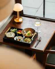 High-angle shot of a complete Japanese bento lunch set served on a tray with a glass of white wine and a modern gold desk lamp on a wooden table near a window. © Simone Oppes