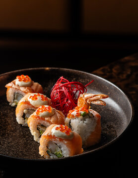Gourmet salmon sushi rolls topped with creamy sauce, red caviar, and ebi tempura shrimp in a dark ceramic dish.