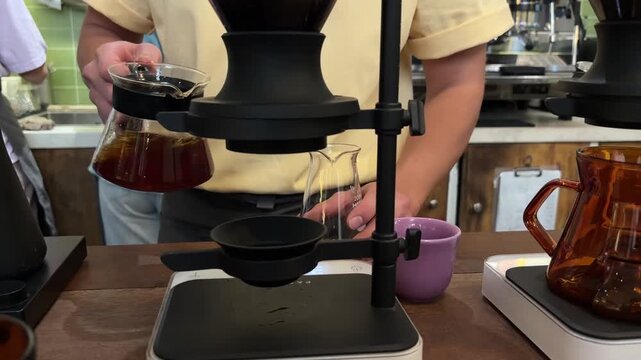 A barista brews coffee using a modern coffee maker. The setup includes a glass carafe, a black stand, and a purple cup. The barista wears a yellow shirt.