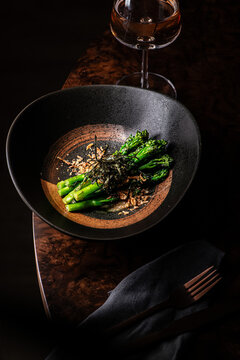 Gourmet sauteed tenderstem broccolini topped with crispy shallots and nori seaweed, paired with a glass of rose wine in a dark setting.