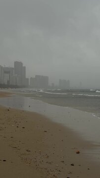 A beach scene with gray skies and rough waves. The shoreline is sandy, and city buildings are visible in the background, shrouded in mist.