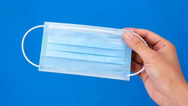 Hand holding blue surgical face mask on blue background, healthcare protection against germs and pollution