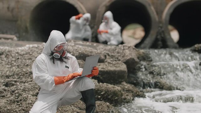 Scientists in hazmat suits inspect polluted water. They record data on laptops near sewage outfalls