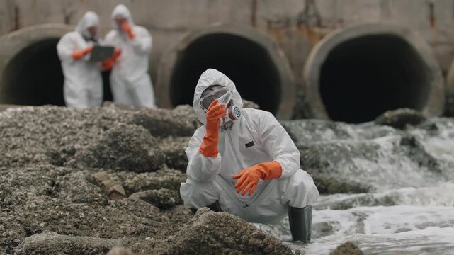 Scientists examine polluted water near industrial pipes. One kneels, testing samples with gloved hands. Others stand guard