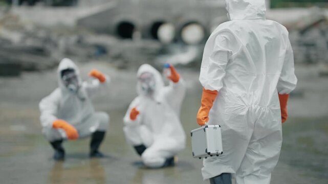 White suits cover workers in muddy, industrial zone. Gloves grip tools and equipment as they move through wet terrain. Faces hidden, focused on task