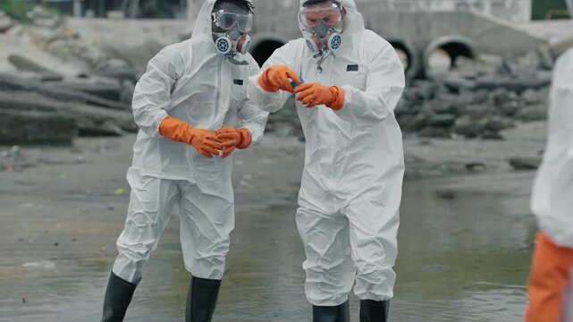 Two scientists in full hazmat suits examine polluted water. They wear masks, gloves, and boots, knee-deep in murky water. One holds a test tube
