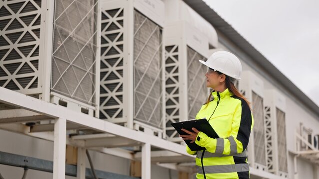 Asian female electrical engineer in safety vest inspecting commercial rooftop cooling tower chillers. Industrial air ventilation system, HVAC maintenance and facility service concept.