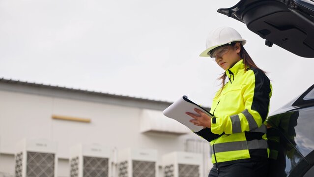 Asian female technician in hardhat checking equipment maintenance checklist on clipboard near cooling units. Commercial HVAC system service, facility engineering and safety concept.