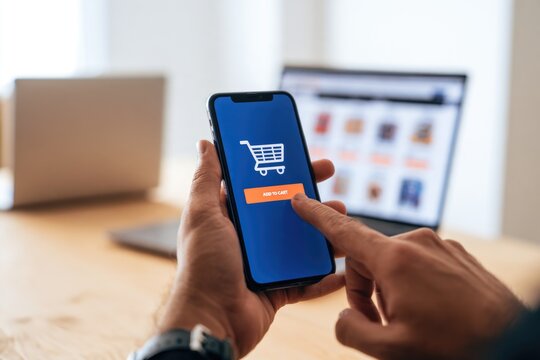 A person uses a smartphone to shop online, tapping the "Add to Cart" button on an e-commerce app with a laptop in the background.