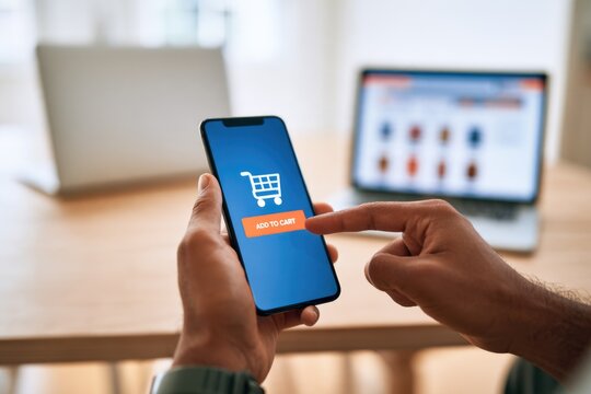 A person uses a smartphone to tap "Add to Cart" on an online shopping app, with a laptop displaying a product page in the background.