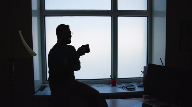 Silhouette of seated bearded man holding mug on windowsill beside bright four-pane frosted window with soft backlit shadows
