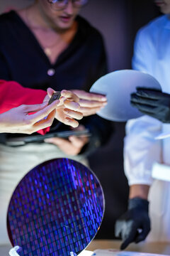 An engineer hands the die to a colleague for a second look while a gloved hand holds a blank disc to one side inside a dimly lit research laboratory late in the working day