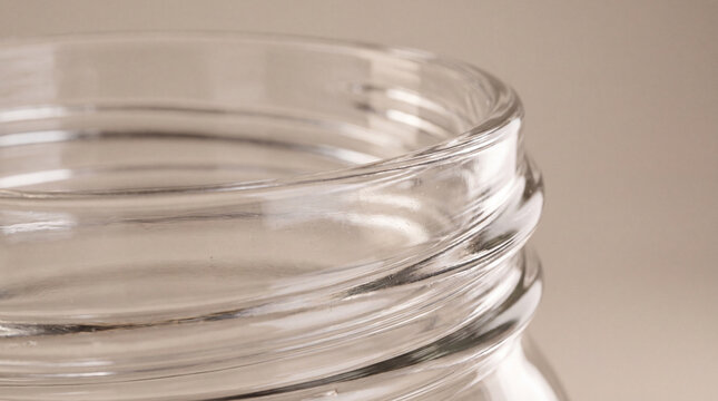 Macro close-up of the rim and threaded neck of a clear glass jar with soft reflections and shallow depth of field on a warm neutral background.
