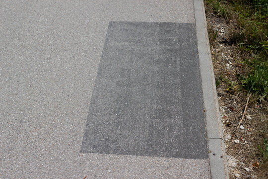 Asphalt pavement with a rectangular patch of darker material indicating recent repair or maintenance work along the roadside near grass and gravel