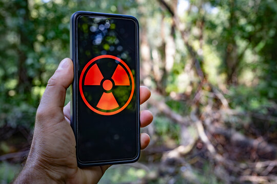 A hand holds a smartphone displaying a red radiation hazard symbol on its screen, set against a natural background.