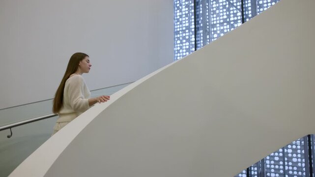 Woman walking up curved modern indoor staircase, hand touching metal handrail, patterned window light glowing behind her in minimalist architecture