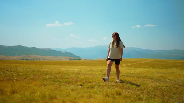 Young woman walking and dancing alone in a wide open grassy field under a clear blue sky, enjoying nature, sunshine, and peaceful mountain landscape views
